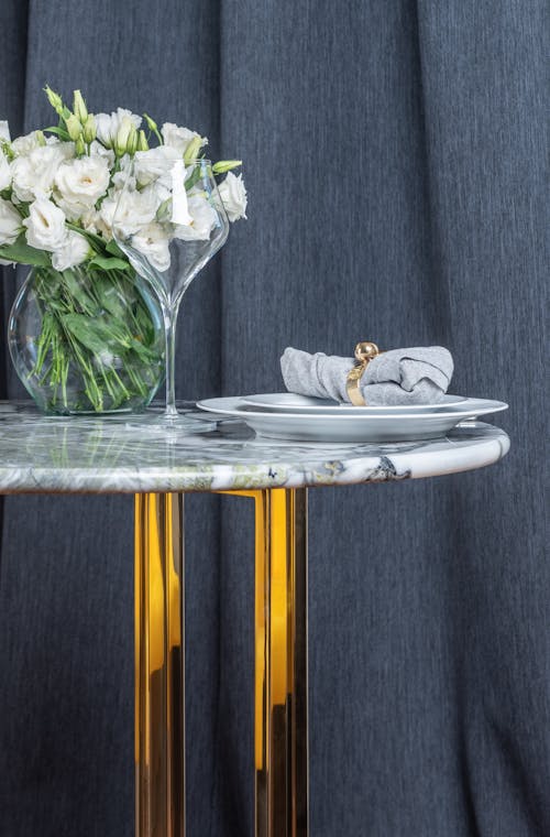 White ceramic plate with crystal wineglass placed on marble table together with bouquet of white flowers in transparent vase