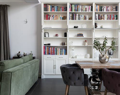 White Wooden Bookcase With Books in the Living Room with Wooden Table, Armchairs and Green Sofa