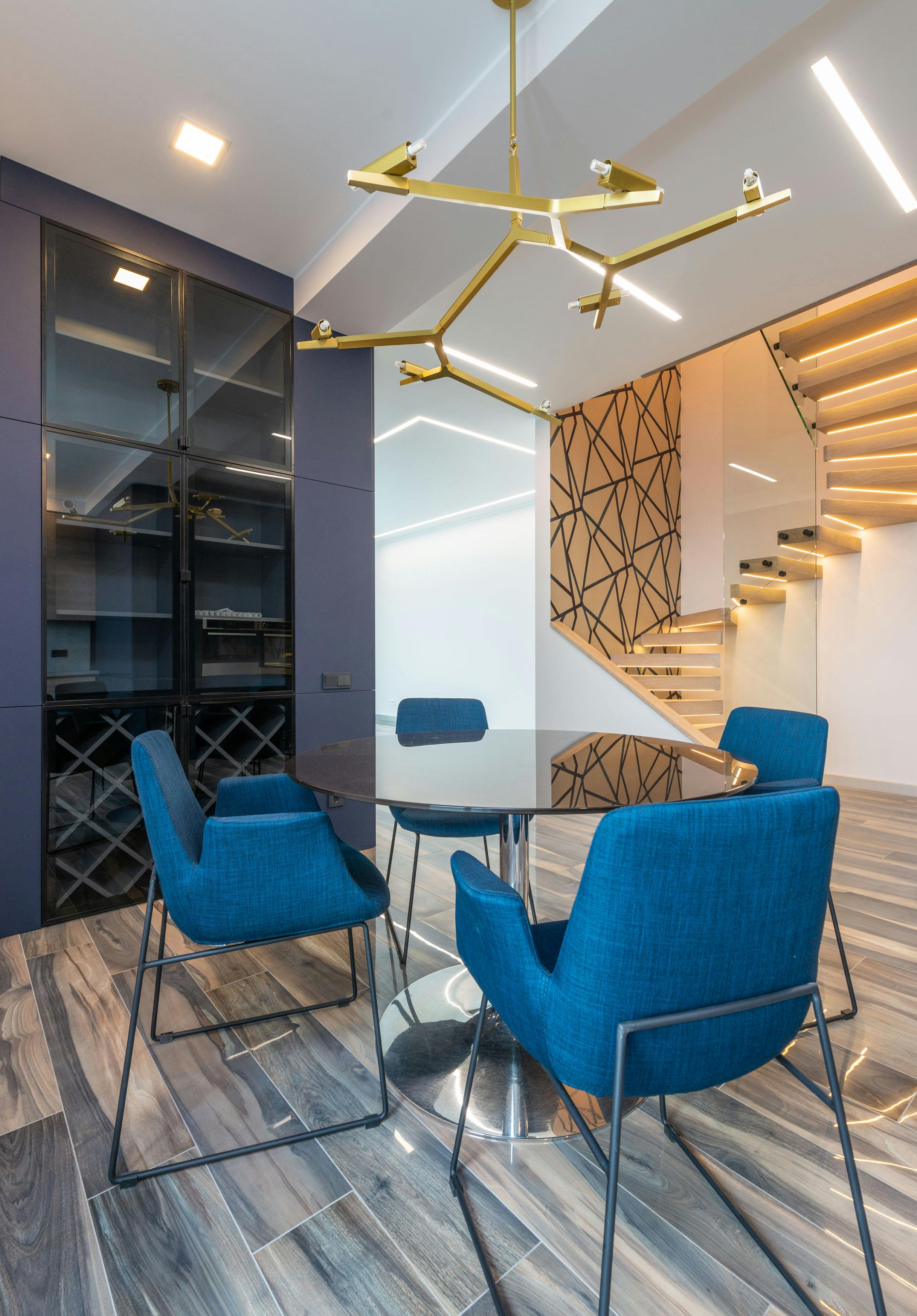 Contemporary dining room highlighted by blue accent chairs, a glass table, and striking overhead lighting.