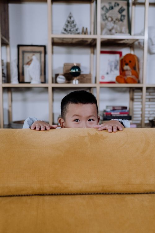 Free A young boy playfully peeks over a mustard yellow couch in a cozy, well-decorated living room. Stock Photo