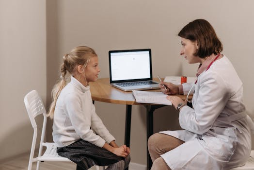 Doctor in lab coat consulting a young girl in a clinic with a laptop on the table.