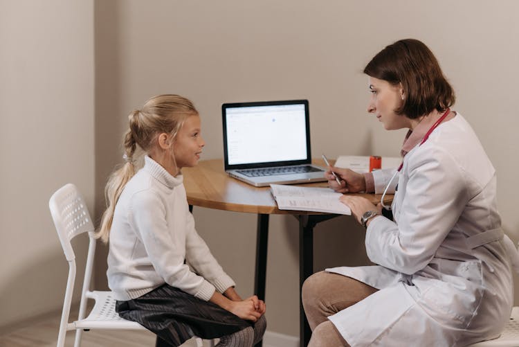 Doctor In White Lab Coat Sitting In Front Of Little Girl And Listening