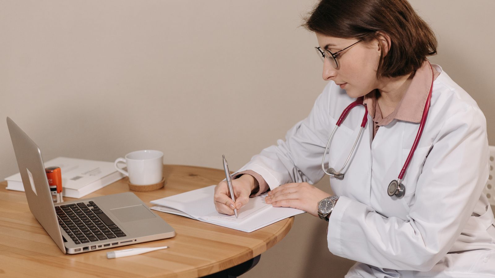 Woman doctor with stethoscope writing at desk with laptop and notes.