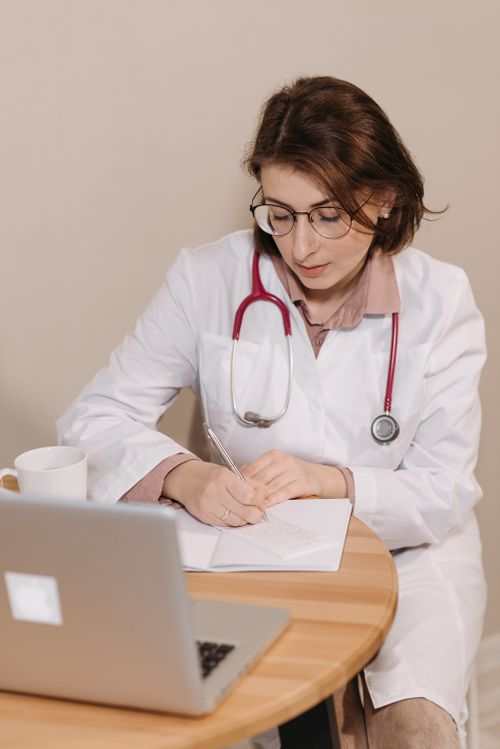 Free Female doctor with stethoscope writing notes in an office setting with laptop. Stock Photo