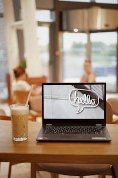 Laptop displaying 'hello' alongside iced coffee on a table in a cafe with blurred background.