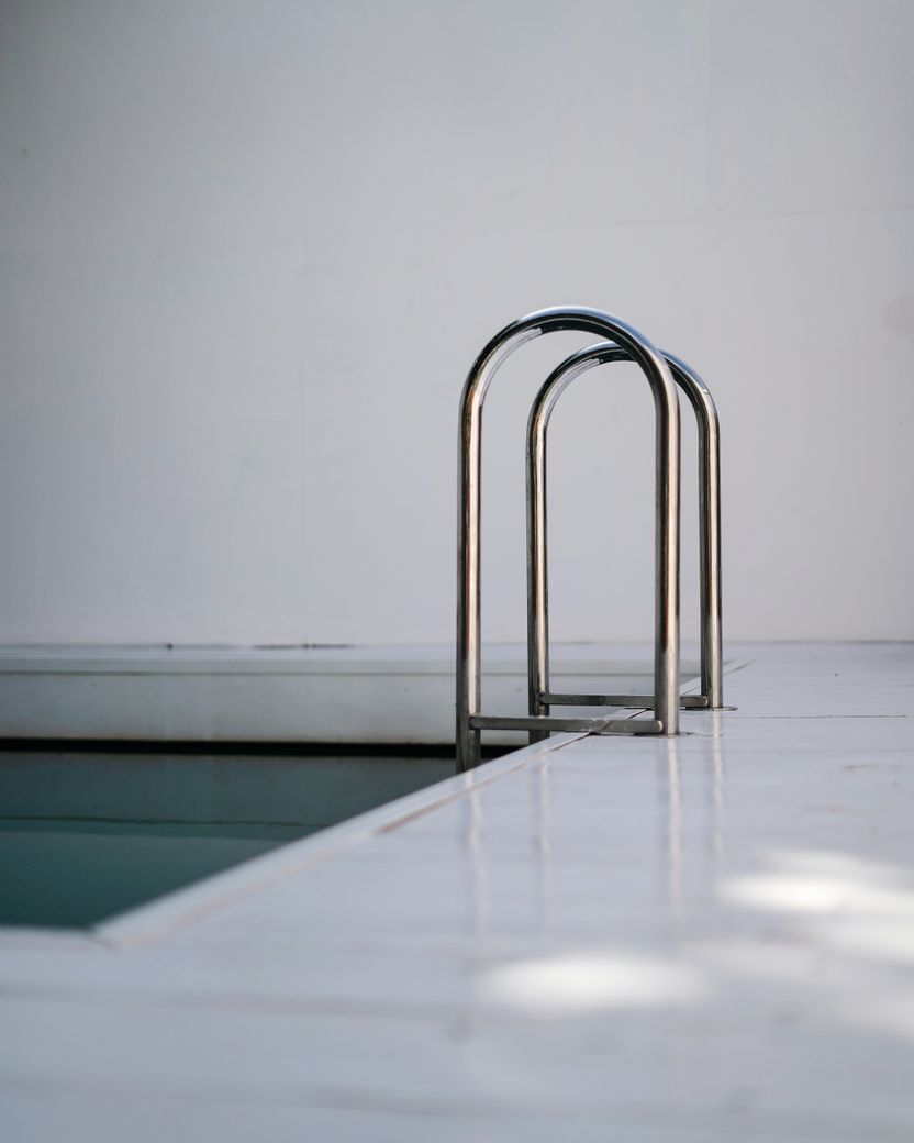 A polished stainless steel handrail of bent tube rising from a pale poolside deck beside still water, against a white wall.