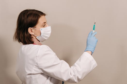Medical practitioner in white coat and face mask preparing a syringe for vaccination indoors.