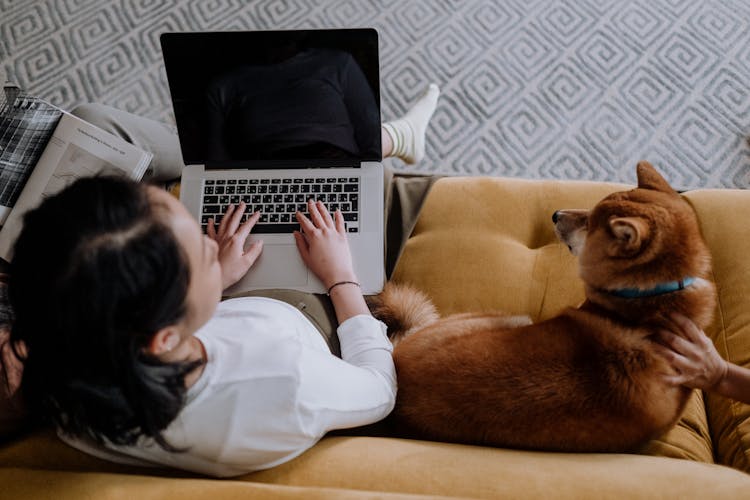 A Woman Using Her Laptop While Sitting On The Couch With Her Dog