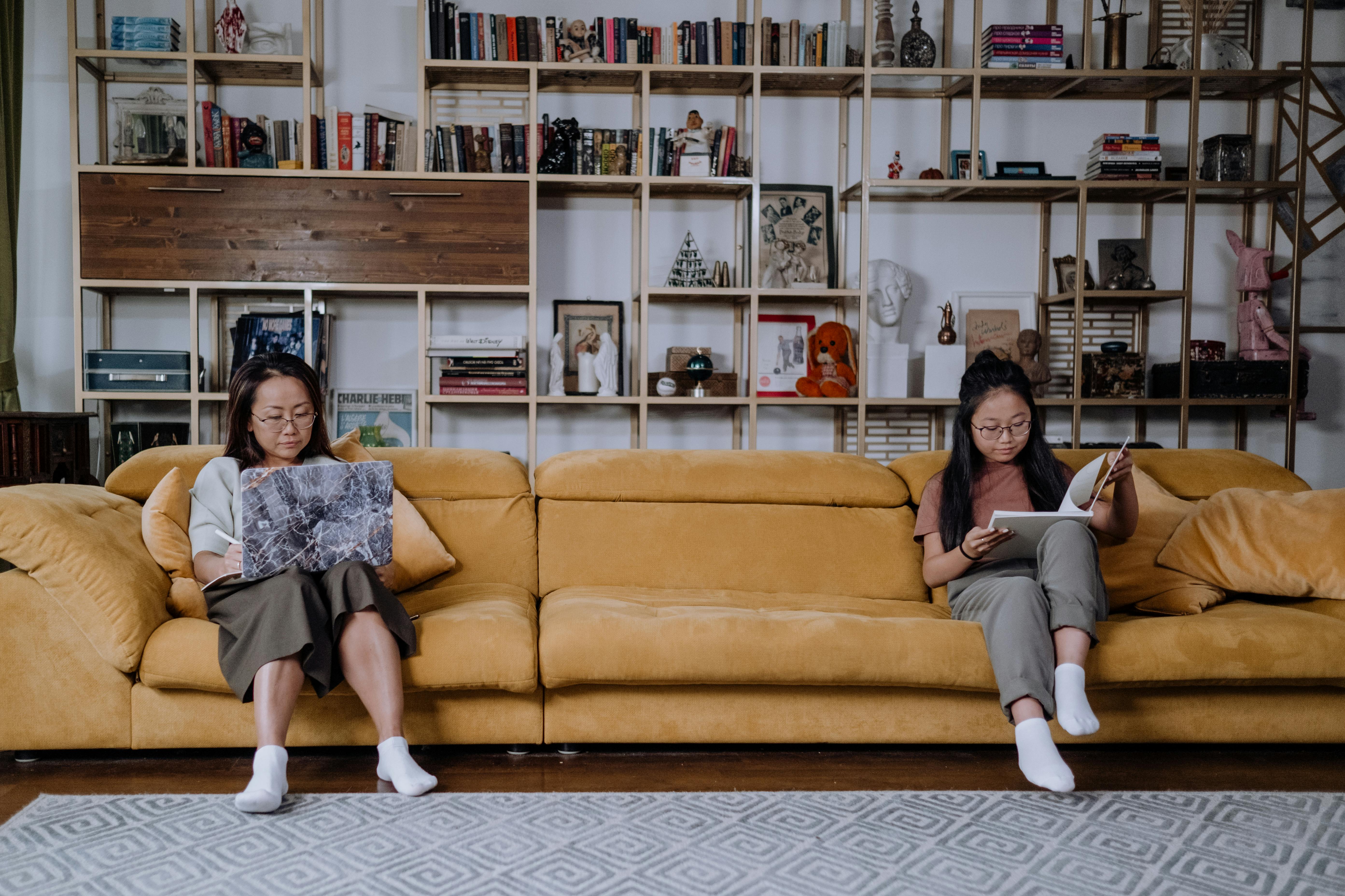 a mother and daughter sitting on the couch while using a laptop and reading a book