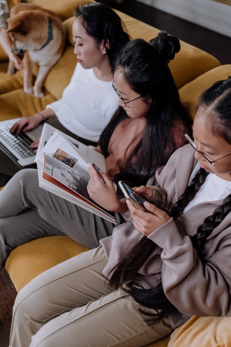 Girls Sitting On The Couch While Using Their Gadgets And Book