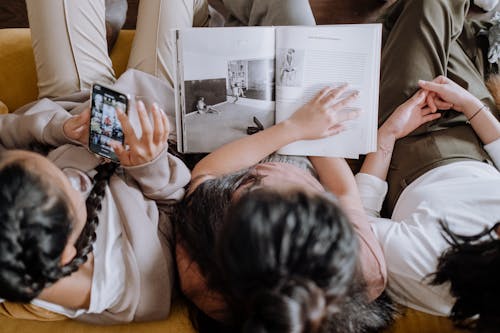 People Sitting on the Couch while Holding a Mobile Phone and a Book