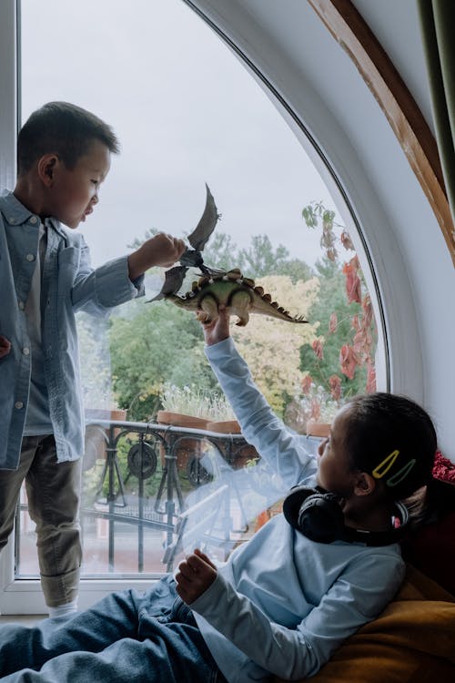 A Young Girl and Boy Holding their Toys while Playing Near the Glass Window