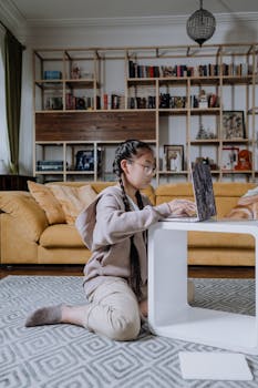 A young girl with braided hair focuses on her laptop, sitting on the floor in a cozy living room.