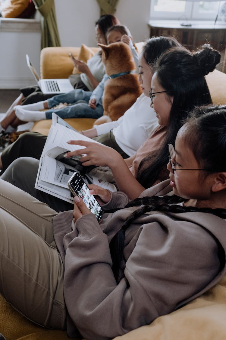 Girls Sitting On The Couch While Reading A Book And Using Mobile Phone