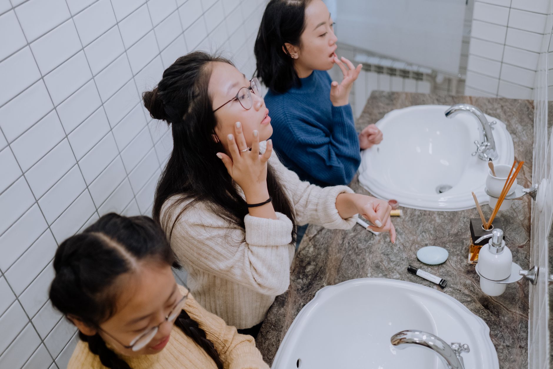 Three teenage girls crowd a bathroom mirror while testing beauty products