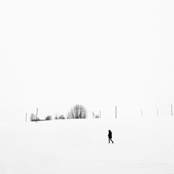 A solitary figure walking across a vast snowy field under a blank sky in winter.