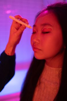 Close-up of an Asian woman receiving makeup with neon pink lighting, focusing on creative eye detail.