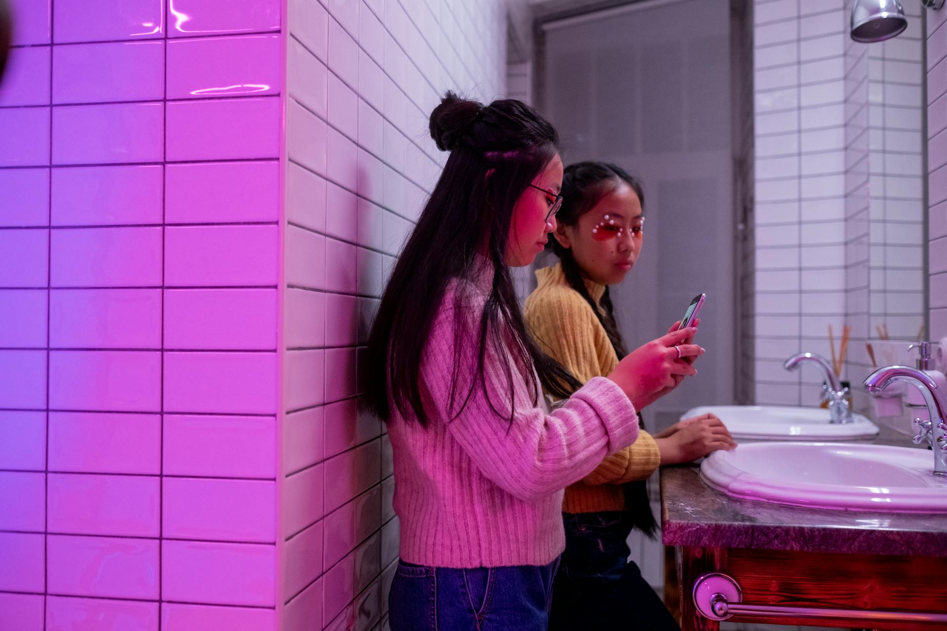 Two young women in a purple-lit bathroom using smartphones near a sink.