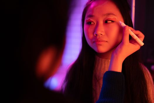 A young woman having makeup applied in a room with vibrant, colorful neon lighting.