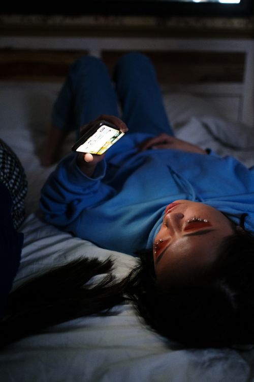 Free A young woman lying on a bed using a smartphone in a dimly lit room, showcasing a relaxed evening vibe. Stock Photo