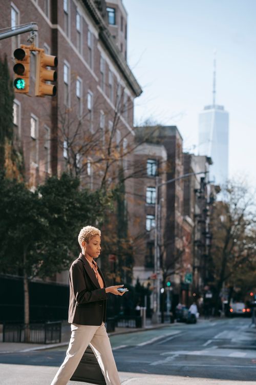 Free Side view of concentrated young African American female entrepreneur with short hair in elegant outfit messaging on smartphone while crossing road in New York Stock Photo