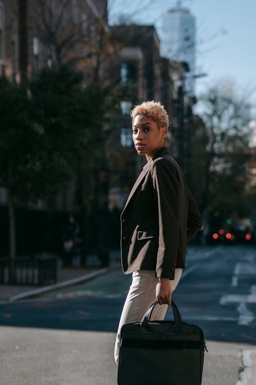 Free Side view of confident young African American female manager with short hair in formal outfit standing on city street with laptop bag in hand and looking at camera Stock Photo