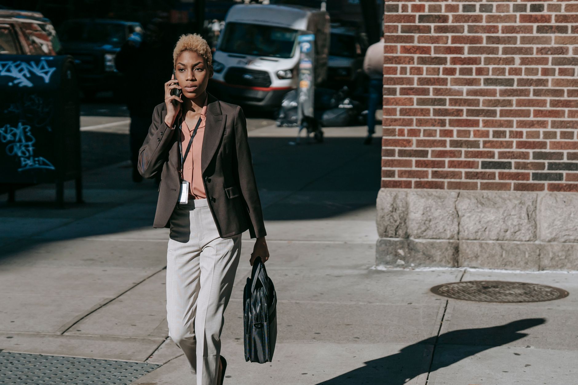 Self assured young African American female manager in stylish formal outfit talking on smartphone while walking on street with laptop bag in hand on sunny day