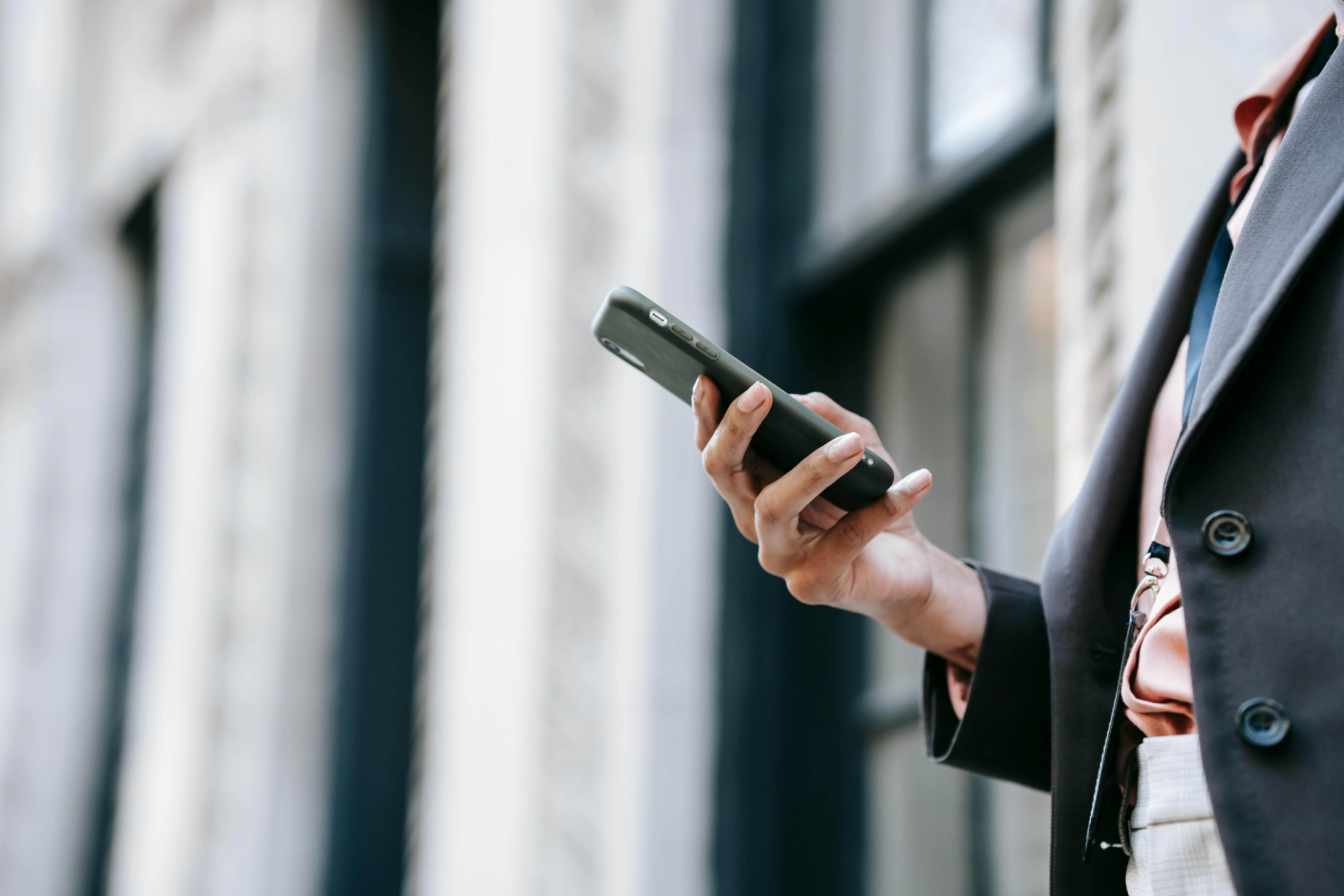 crop businesswoman messaging on smartphone while standing near building on street