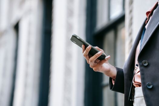 Side view of crop unrecognizable female manager in stylish outfit using mobile phone while standing on street near building before going to work