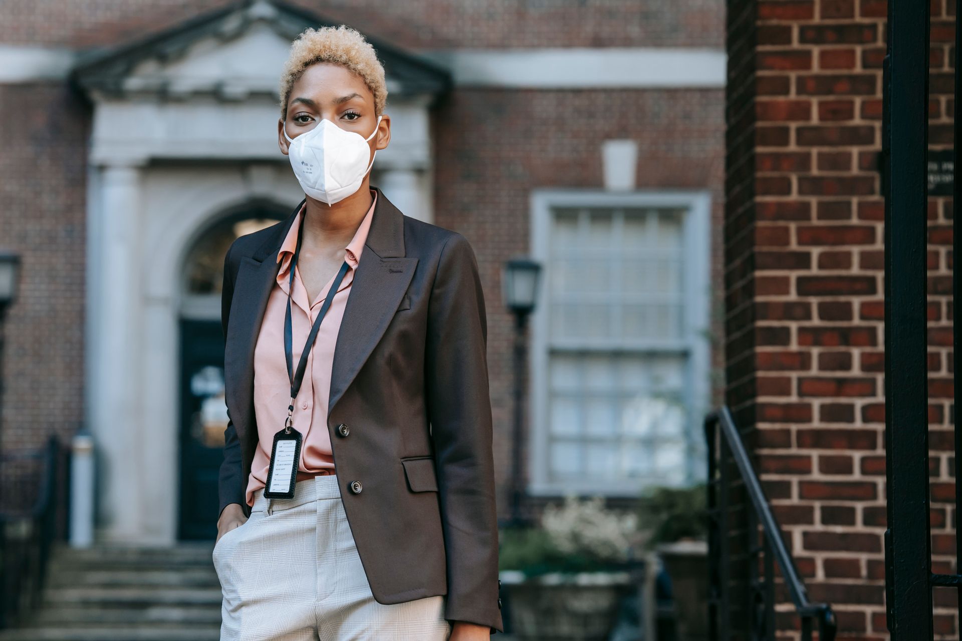 Serious young black female entrepreneur with short dyed hair in stylish suit and protective mask standing on street near brick building with had in pocket and looking at camera
