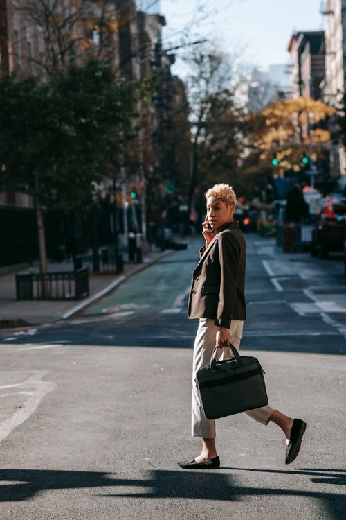 Free Full body African American businesswoman in black jacket with laptop bag talking on smartphone while crossing road and looking at camera Stock Photo