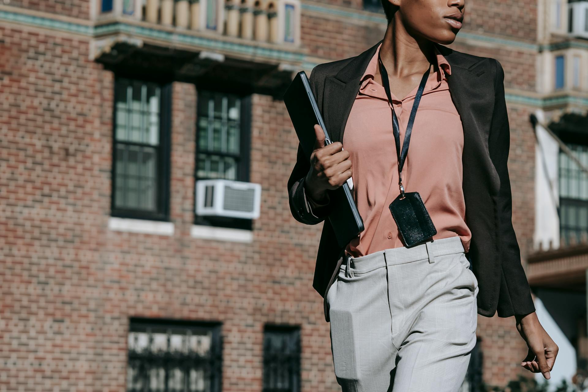 Crop anonymous serious African American female entrepreneur in classy outfit with badge walking with laptop and smartphone in pocket