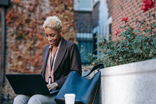 Smiling young African American female freelancer in stylish jacket with badge working on laptop while sitting on bench near flowerbed