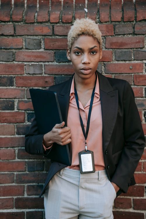 Free Confident young African American female entrepreneur in stylish jacket with badge and laptop standing near brick wall with hand in pocket and looking at camera Stock Photo