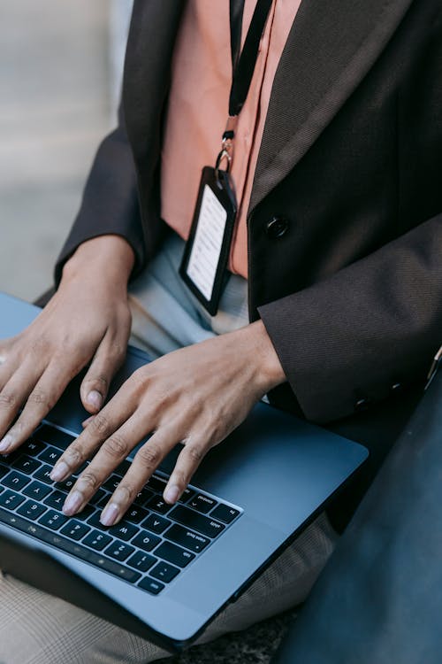 From above of crop faceless female freelancer in black jacket with badge typing on laptop while working on new project