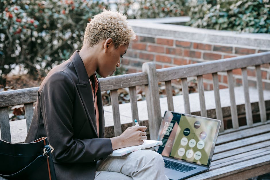 High angle side view of focused African American female employee writing notes while watching presentation on laptop sitting on wooden bench