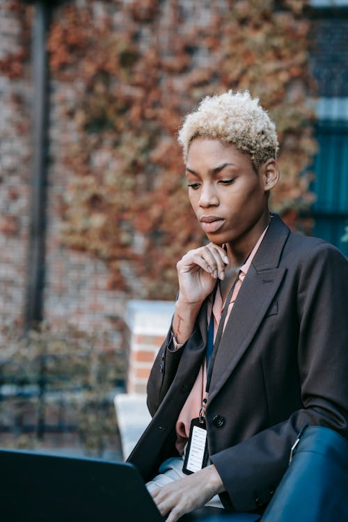 Pensive young African American female freelancer in formal jacket with badge working on laptop touching chin