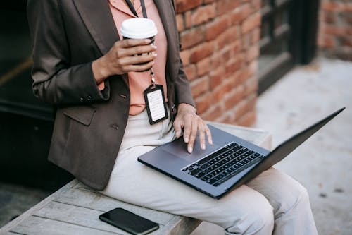 From above of crop anonymous ethnic female manager with name tag using touchpad of laptop with cup of hot drink