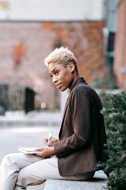 Side view of confident African American female employee in formal clothes sitting on bench in park and writing in notepad