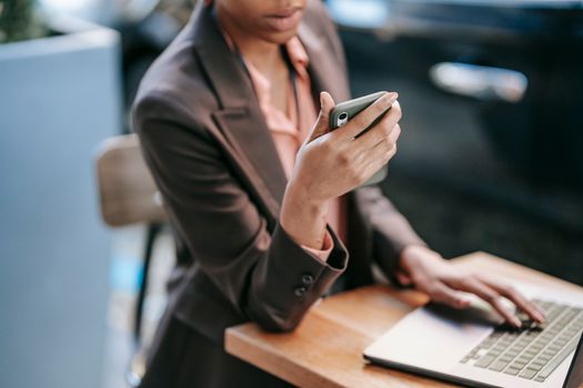 High angle of crop African American female employee wearing formal clothes typing on laptop while checking message on cellphone