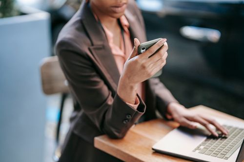 Free High angle of crop African American female employee wearing formal clothes typing on laptop while checking message on cellphone Stock Photo