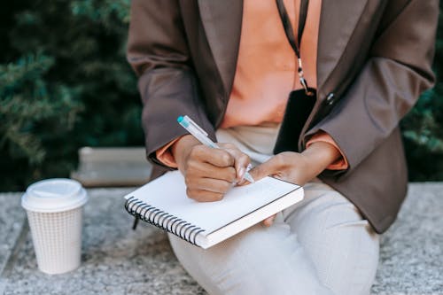 From above of crop anonymous female employee wearing formal clothes writing notes in notebook while sitting with hot drink in city park