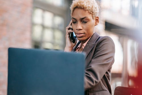 Serious black woman calling on smartphone while watching laptop