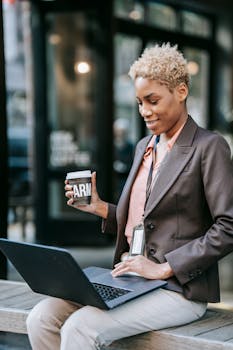 Businesswoman sitting outdoors with a laptop, enjoying coffee break.