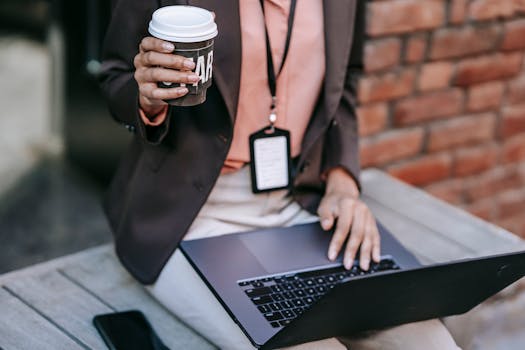 Adult woman multitasking outdoors with a laptop and coffee cup, symbolizing remote work.