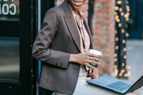 Free Crop anonymous African American female entrepreneur in classy jacket with badge using laptop and drinking coffee to go Stock Photo