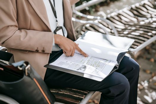 From above crop faceless female student in formal jacket sitting on bench in park and reading texts printed on paper