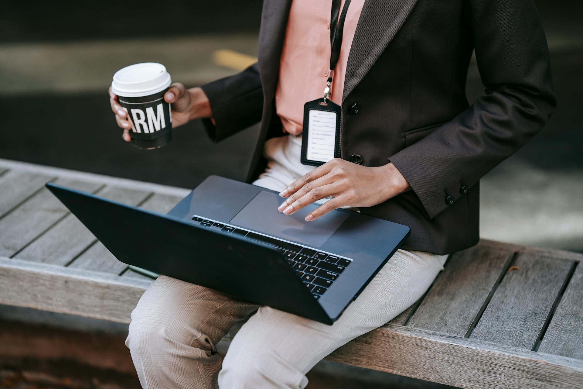 From above of crop anonymous female entrepreneur in stylish black jacket with badge sitting on wooden bench drinking coffee and using laptop