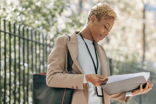 Free Glad young African American female entrepreneur in stylish jacket with badge and bag reading documents Stock Photo
