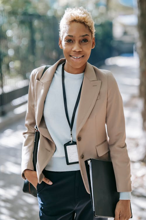 Content young African American female entrepreneur in formal jacket with hand in pocket and folder with documents looking at camera